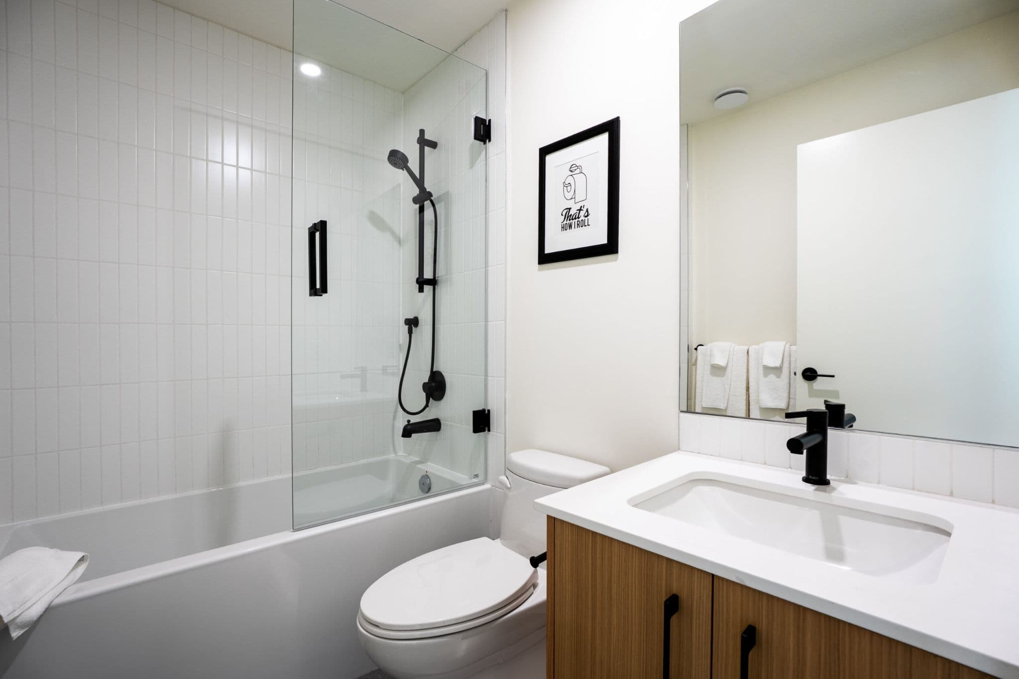 The Linden Townhomes – A bathroom with a soaker tub behind a glass shower panel, black fixtures, a wall-mounted toilet, and a wood vanity with a white countertop and undermount sink.