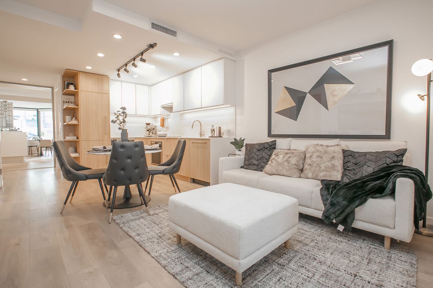 Elements – Furnished show-home living and dining area with a round dining table, dark tufted chairs, a white sofa with faux-fur pillows, and a geometric wall art piece.