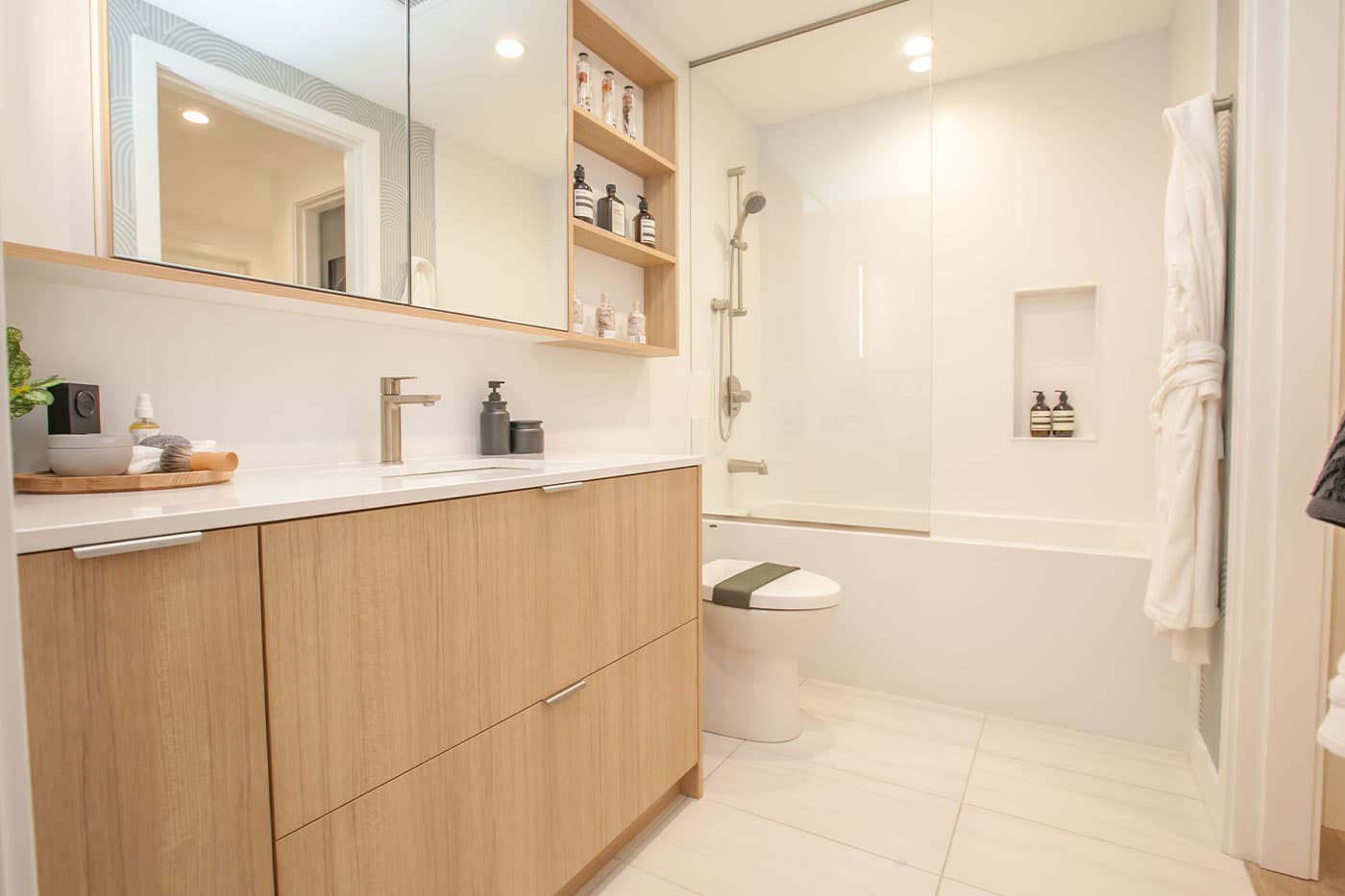 Elements – Bathroom with a wood vanity, large mirror, open shelving with toiletries, a toilet, and a glass-enclosed tub/shower combo.