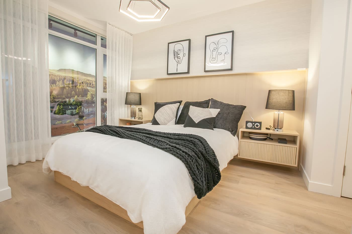 Elements – Bedroom with a white bedspread, dark knit throw, nightstands with lamps, line-art portraits above the headboard, and a floor-to-ceiling window showing a hillside view.