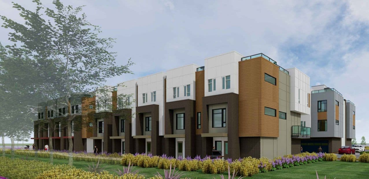 Yorkson SW Townhomes – A row of modern three-story townhomes features a mix of dark brick facades, warm wood-toned siding, and white upper-level paneling. Low flowering bushes line the manicured front lawn under a bright, partly cloudy sky.