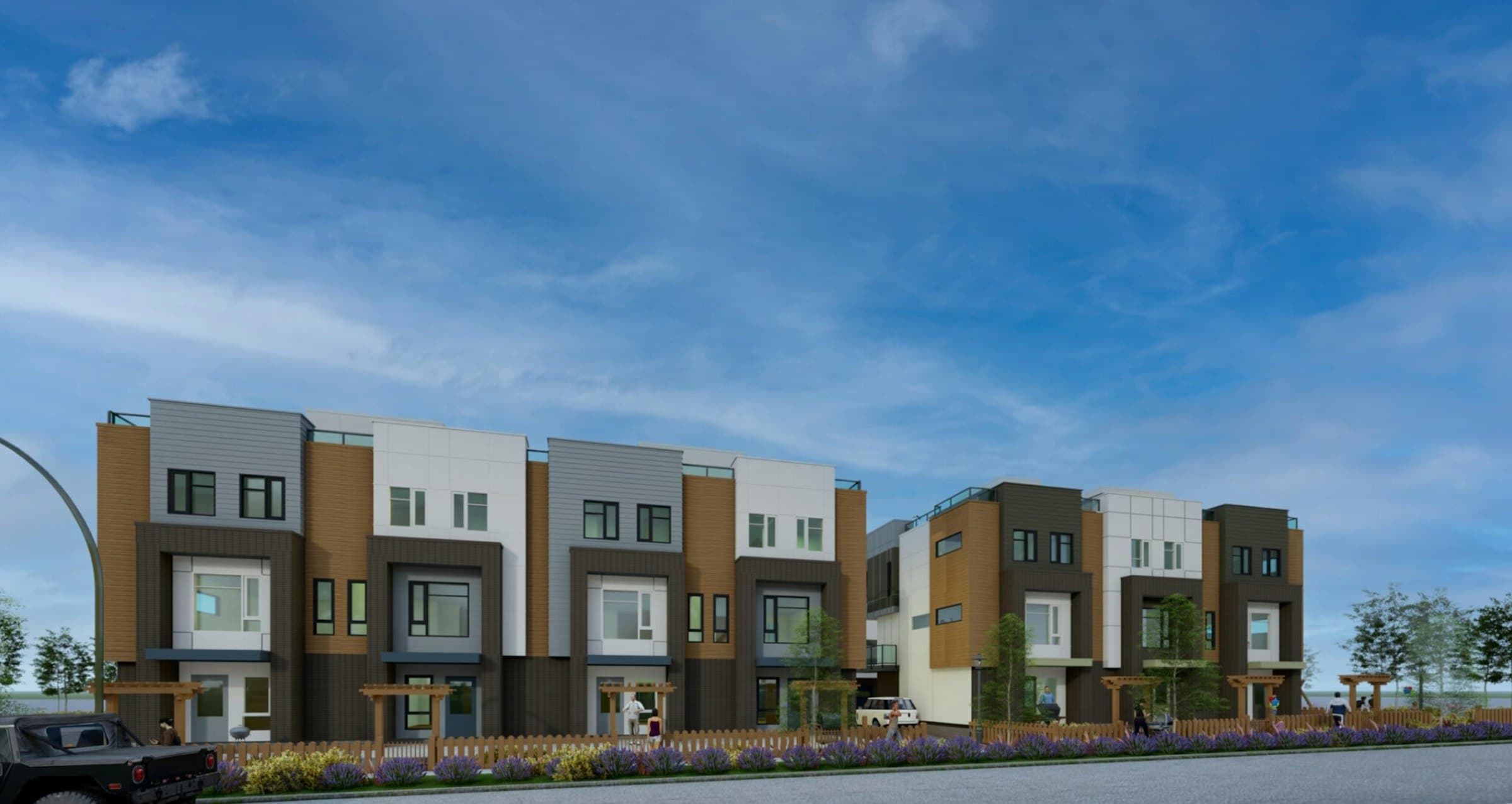 Yorkson SW Townhomes – A street-level architectural rendering shows a row of modern three-story townhomes featuring a mix of dark brick, warm wood-look siding, grey horizontal lap siding, and white panels. Wooden fences, small arbors, and purple floral landscaping line the paved street in front under a clear blue sky.