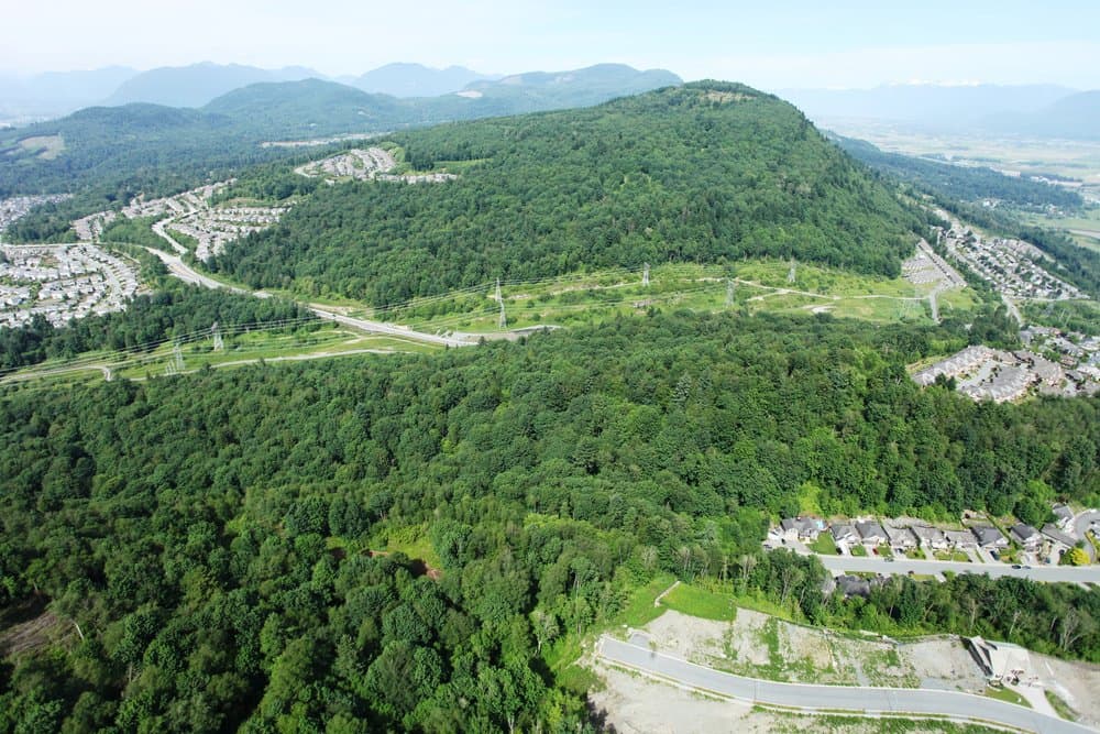 Vicarro – Aerial drone view of a forested hill with power line corridors, surrounded by residential subdivisions and mountains.