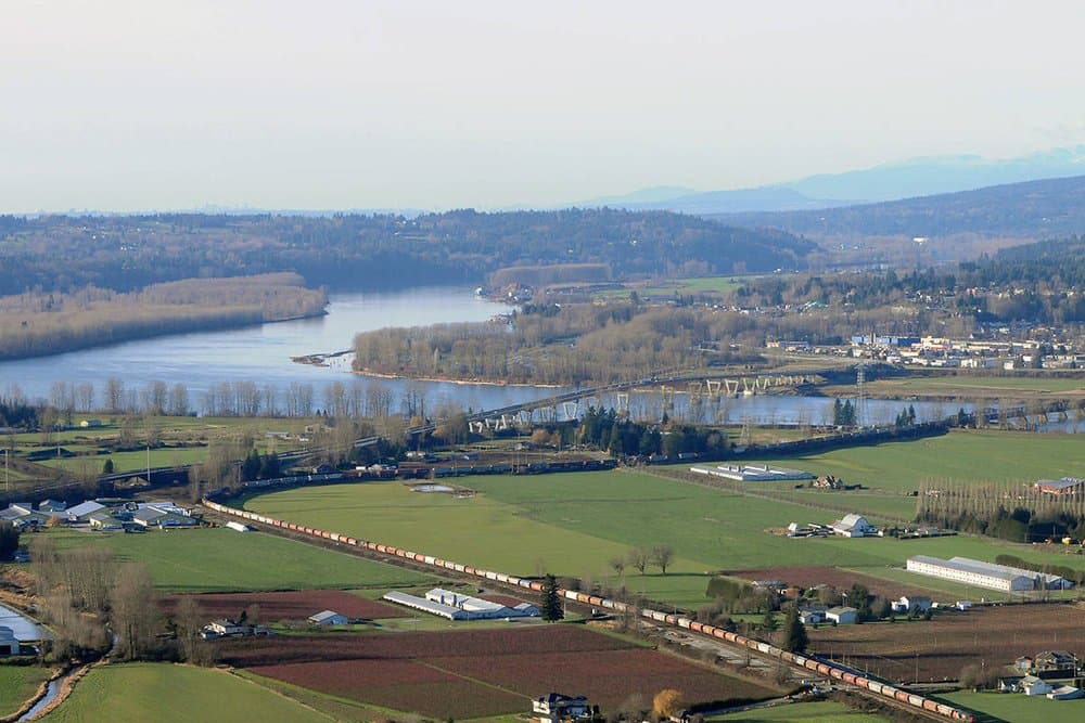 Vicarro – Aerial view of a river valley with a bridge, rail line, and farmland, mountains visible in the haze.