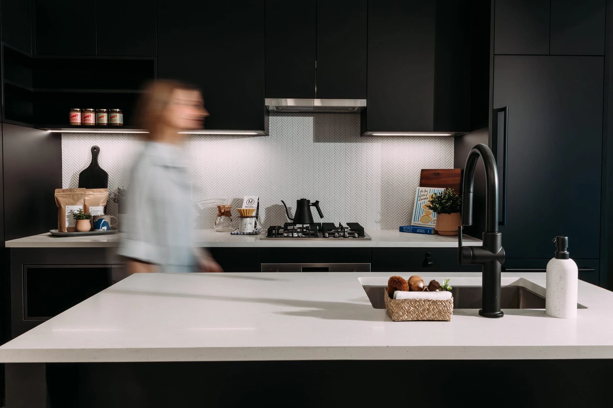 Ace on the Drive – Dark matte black kitchen with gas cooktop, black faucet at the island sink, herringbone tile backsplash, and a person walking blurred through the space.