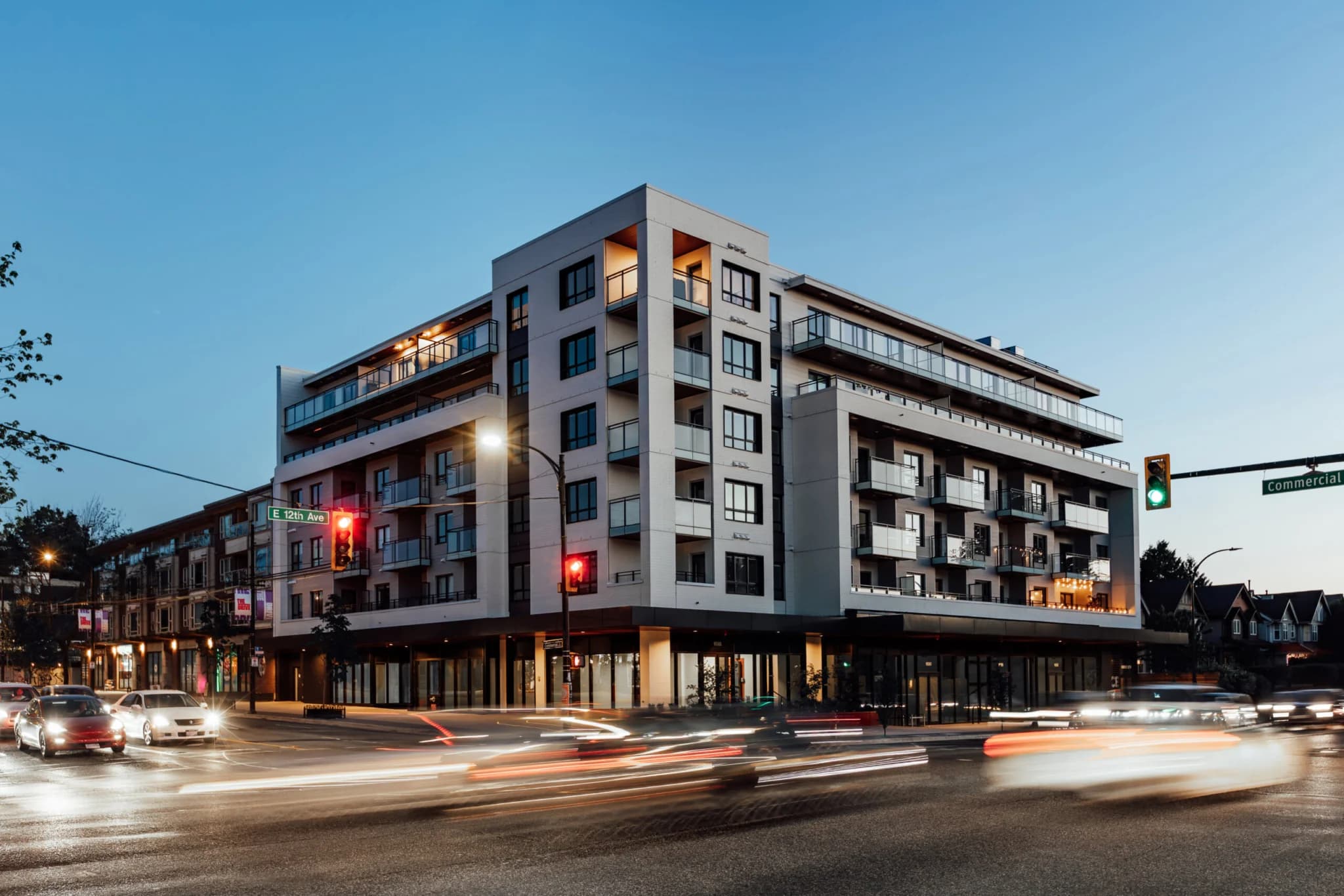 Ace on the Drive – Night photo of the building's corner at E 12th Ave and Commercial, with lit balconies, ground-floor retail, and light-trail traffic passing.