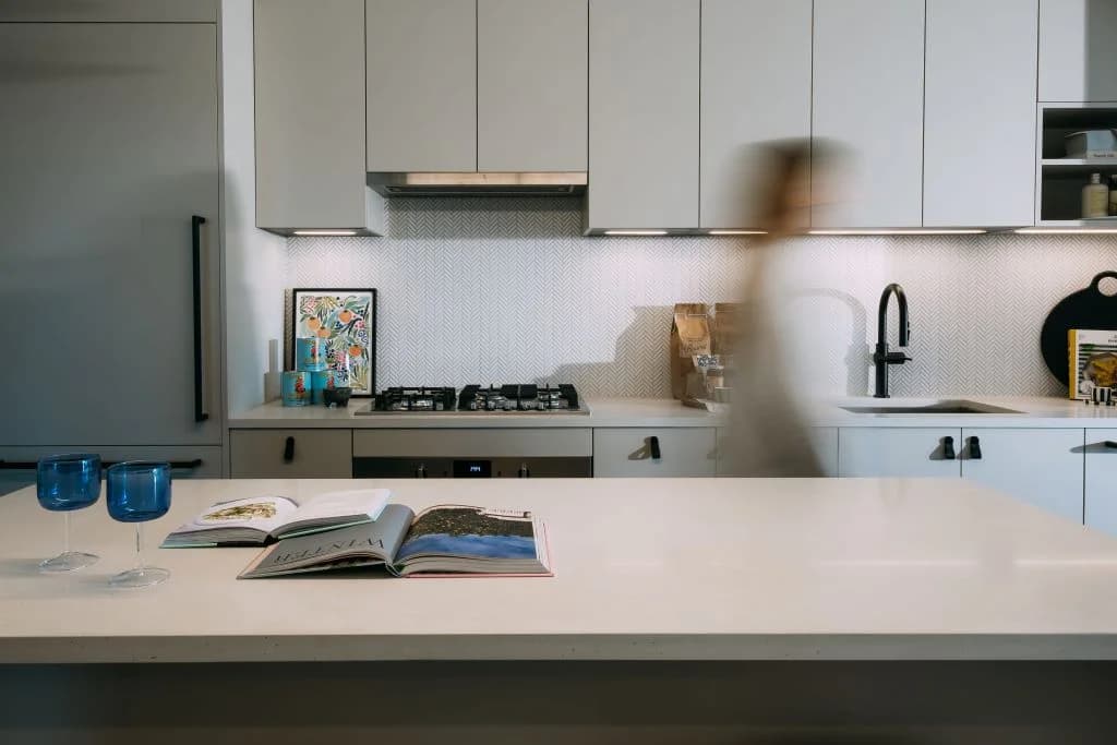Ace on the Drive – Kitchen island countertop with wine glasses and open books, light grey cabinetry and herringbone tile backsplash behind, a person walking blurred in the background.