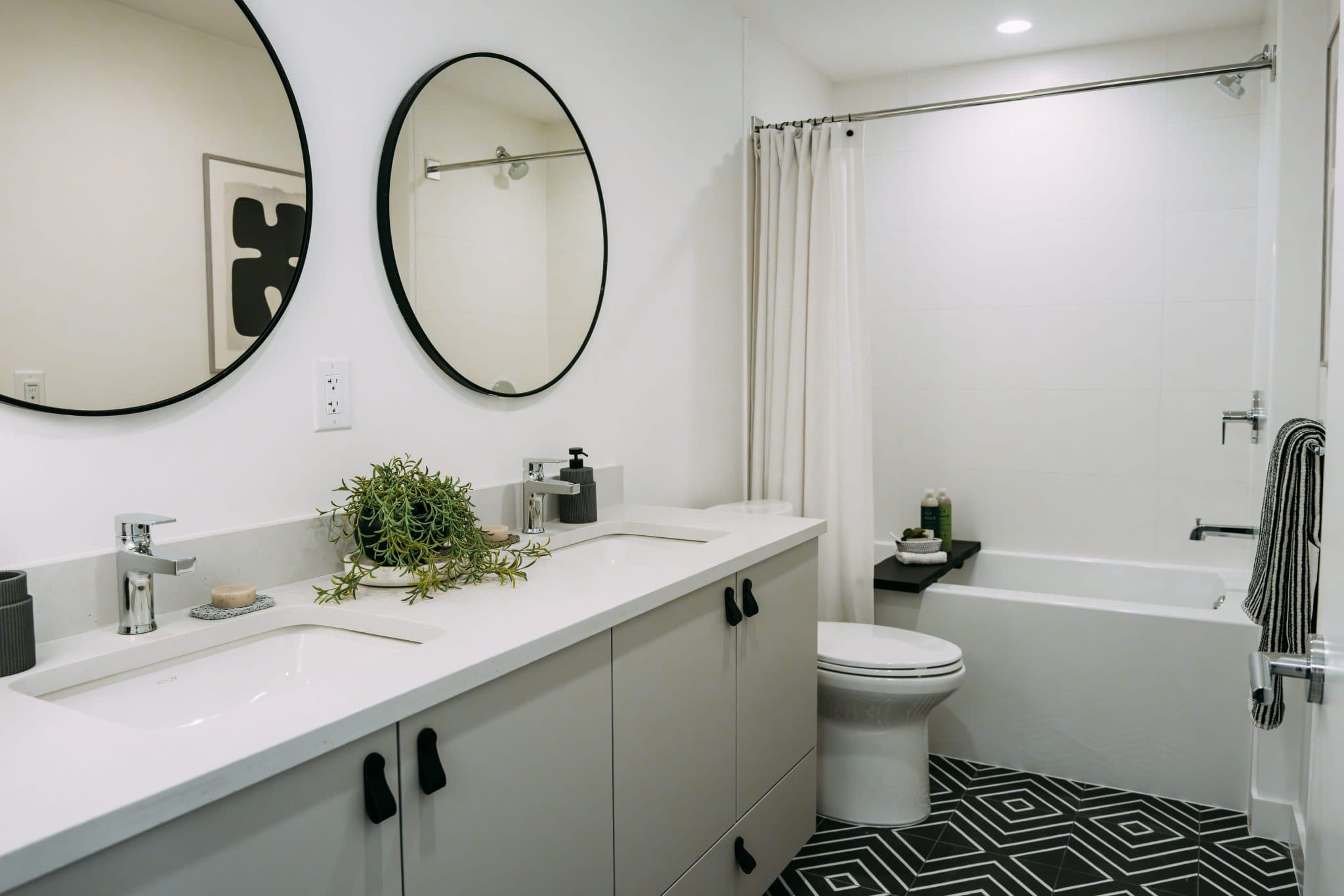 Ace on the Drive – Bathroom with a long white double-sink vanity, two round mirrors, and a separate bathtub with shower curtain and black-and-white patterned floor tile.