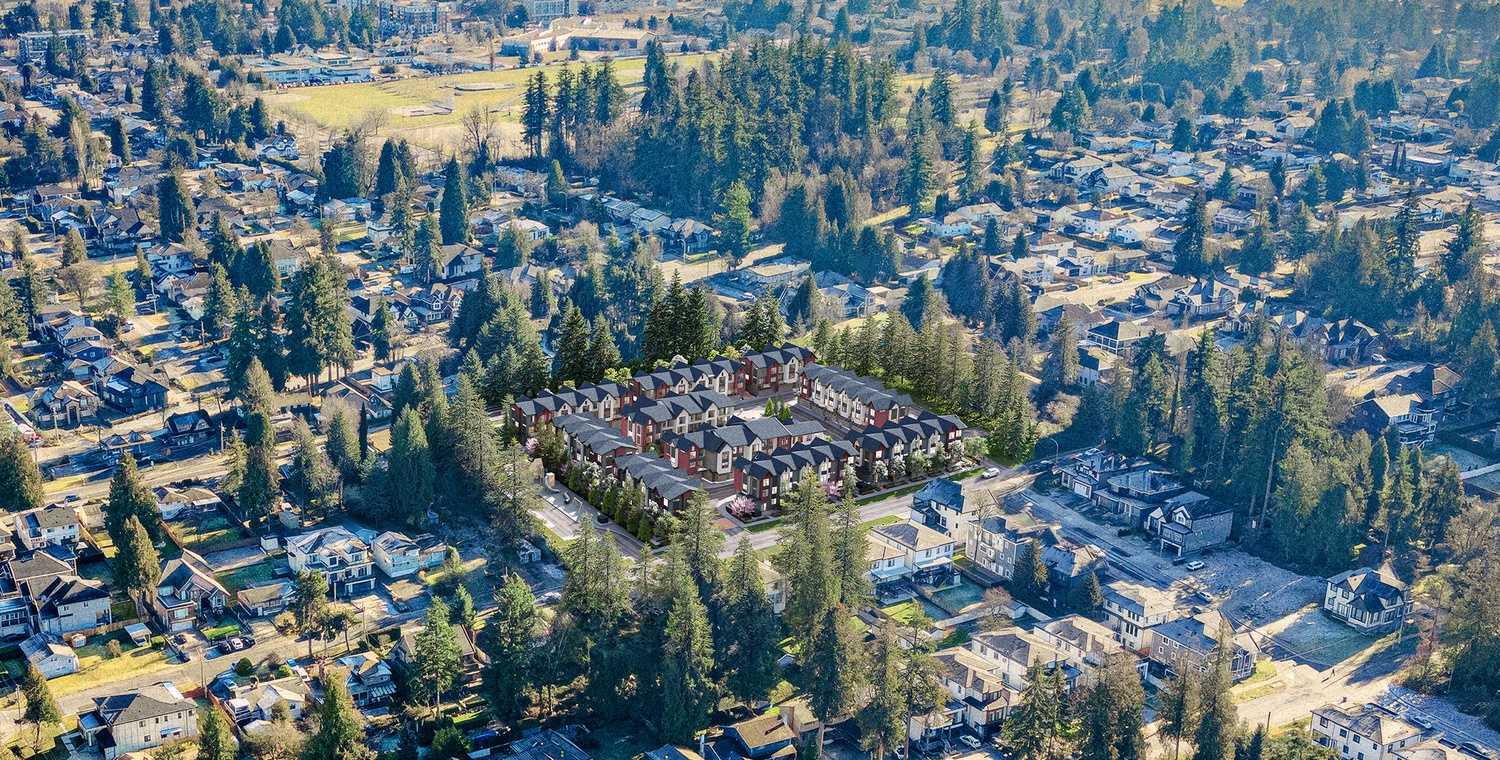 The Westminster – A high-angle drone view showcases a newly rendered townhouse community nestled within a heavily wooded, sprawling suburban neighborhood. The complex features multiple peaked-roof buildings with dark and reddish siding, completely surrounded by mature evergreen trees, residential streets, and single-family homes under bright daylight.