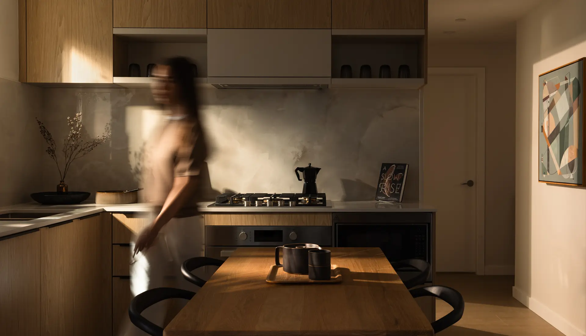 Elmwood – A contemporary kitchen and dining space with natural wood cabinetry, a light marble-patterned backsplash, and a wooden dining table flanked by black chairs. Warm, dramatic sunlight casts deep shadows across the room as a blurred person walks through the space.