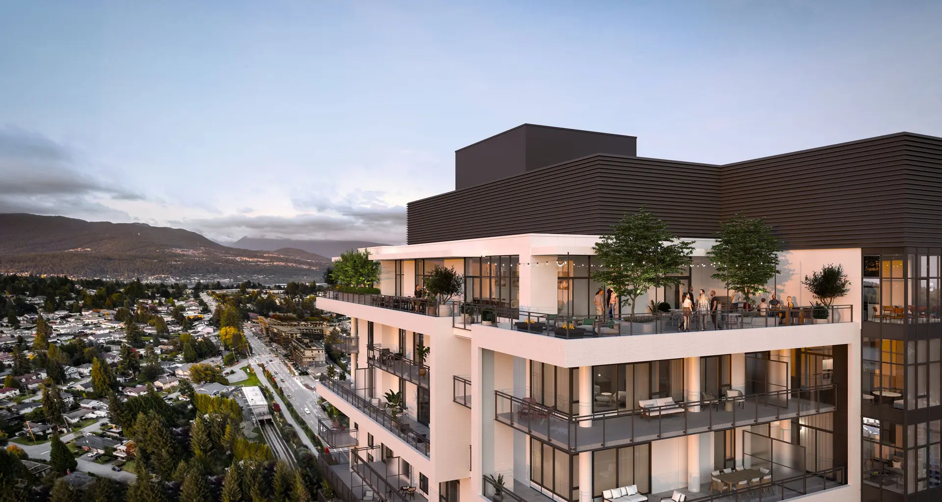 Elmwood – An elevated view of a contemporary mid-rise residential building featuring spacious private balconies and a top-floor terrace where people gather among potted trees and string lights. In the background, a suburban neighborhood and roadway stretch toward forested mountains beneath an early evening sky.