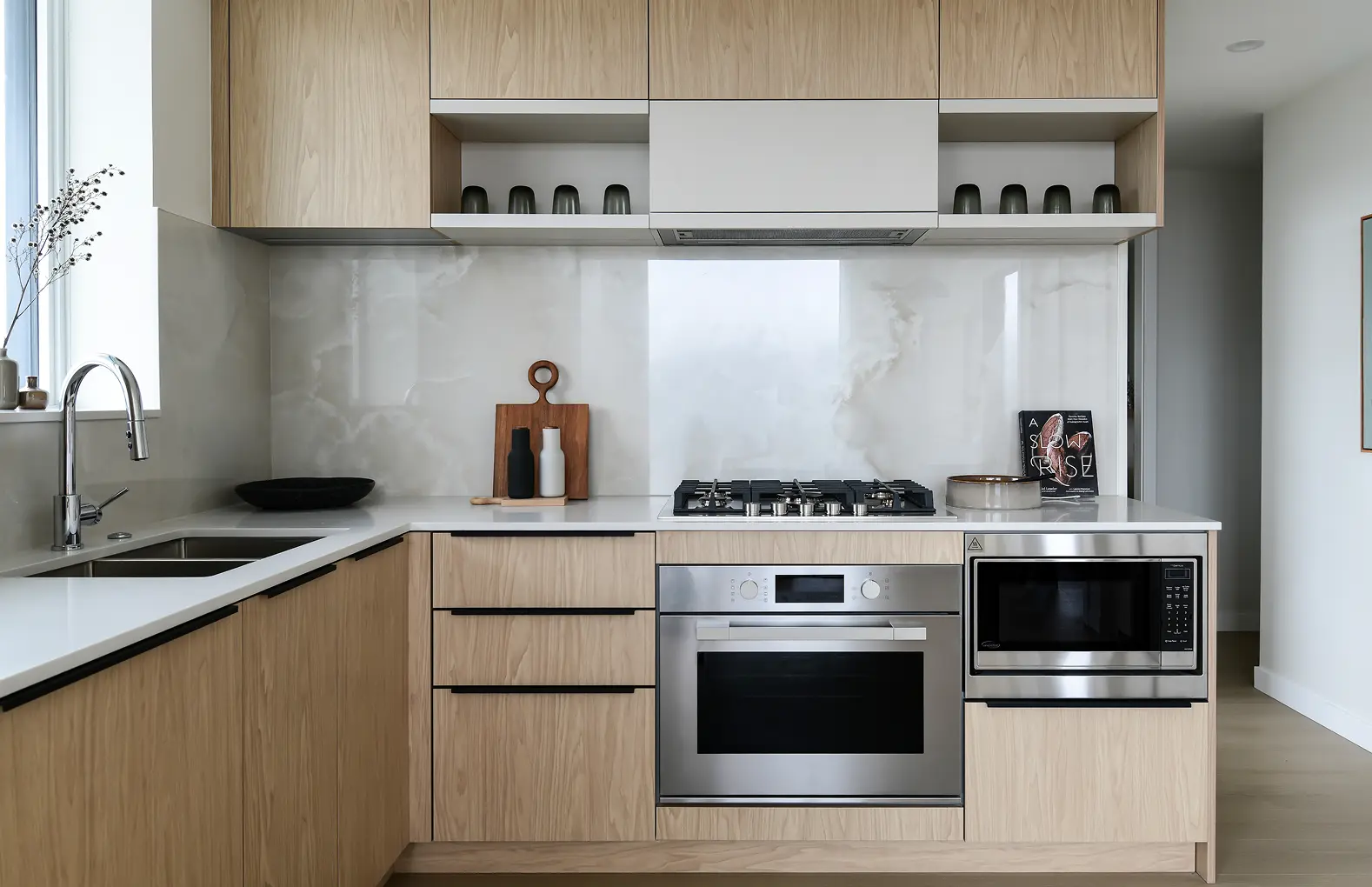 Elmwood – A modern, clean kitchen featuring light natural wood flat-panel cabinetry, white quartz countertops, and a polished slab backsplash with subtle marble veining. Built-in stainless steel appliances, including a gas cooktop, under-counter oven, and microwave, are seamlessly integrated alongside an undermount sink with a gooseneck faucet next to a bright window.