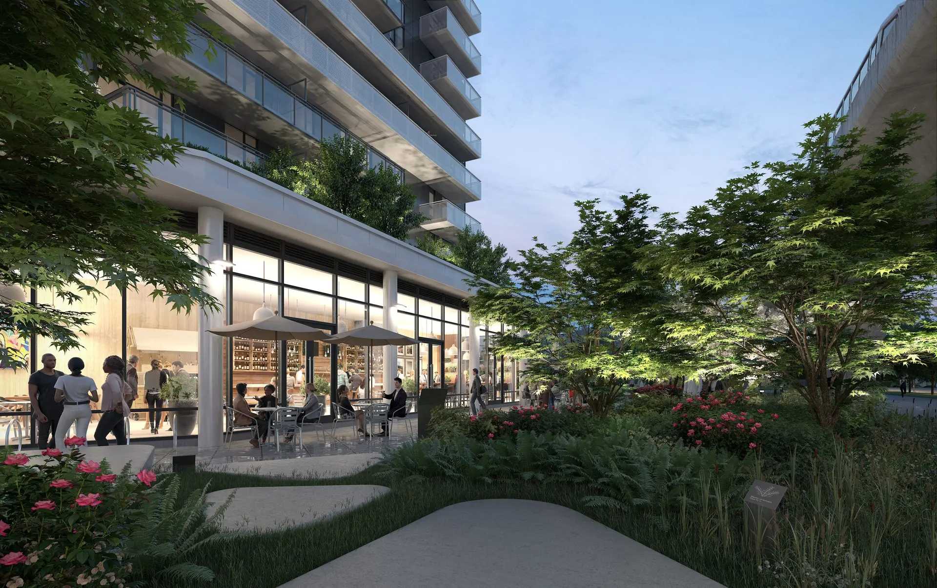 Elmwood – A ground-level view of a modern residential building featuring expansive floor-to-ceiling glass commercial spaces with people gathered at outdoor cafe seating under umbrellas. Lush landscaping with leafy trees, ferns, and pink flowers surrounds winding concrete walkways in the foreground during early evening.