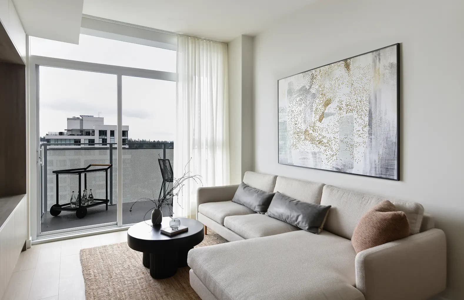 Elmwood – A bright, contemporary living room features a cream-colored sectional sofa, a textured rug, and a low black coffee table beneath a large abstract canvas with gold flecks. Floor-to-ceiling sliding glass doors open onto a private balcony furnished with a bar cart and chair, looking out toward neighboring buildings under an overcast sky.