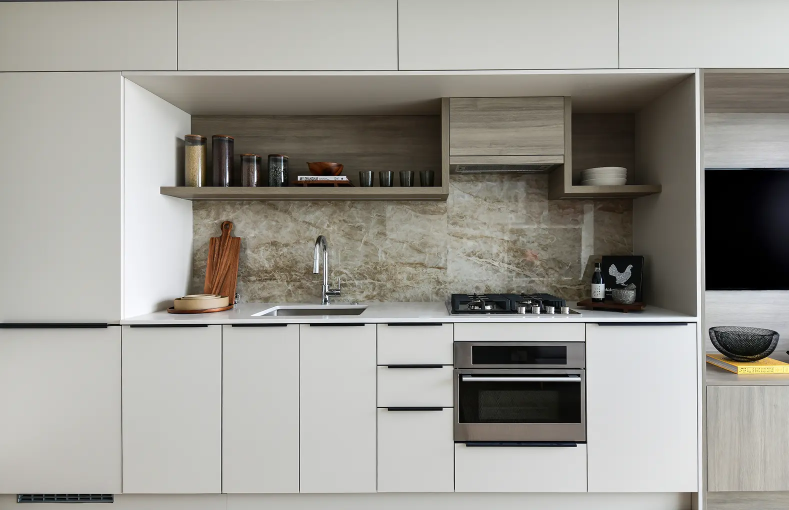 Elmwood – A modern minimalist kitchen featuring flat-panel white cabinetry, a built-in stainless steel oven, and an integrated gas cooktop. The counter space is complemented by a natural stone slab backsplash, a chrome faucet, and open wooden upper shelving styled with jars and dishware.