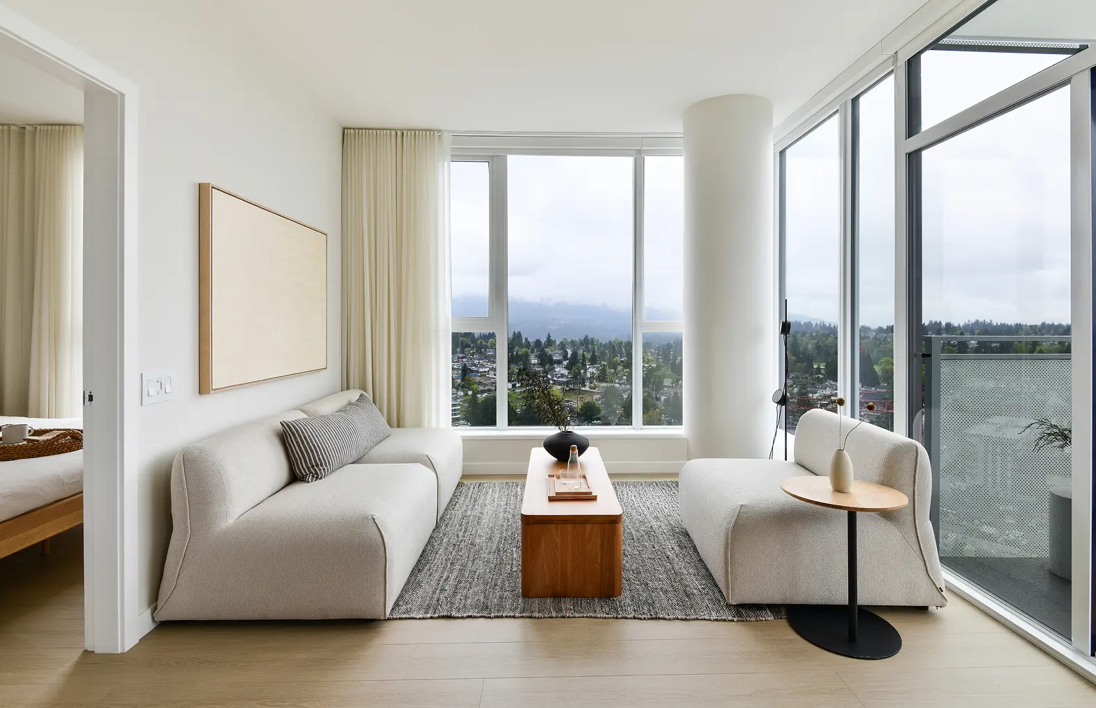Elmwood – A bright, minimalist apartment living room features light neutral upholstered seating, a warm wood coffee table, and light wood flooring. Large floor-to-ceiling corner windows and a glass balcony door look out over a lush green neighbourhood and misty distant mountains under an overcast sky.