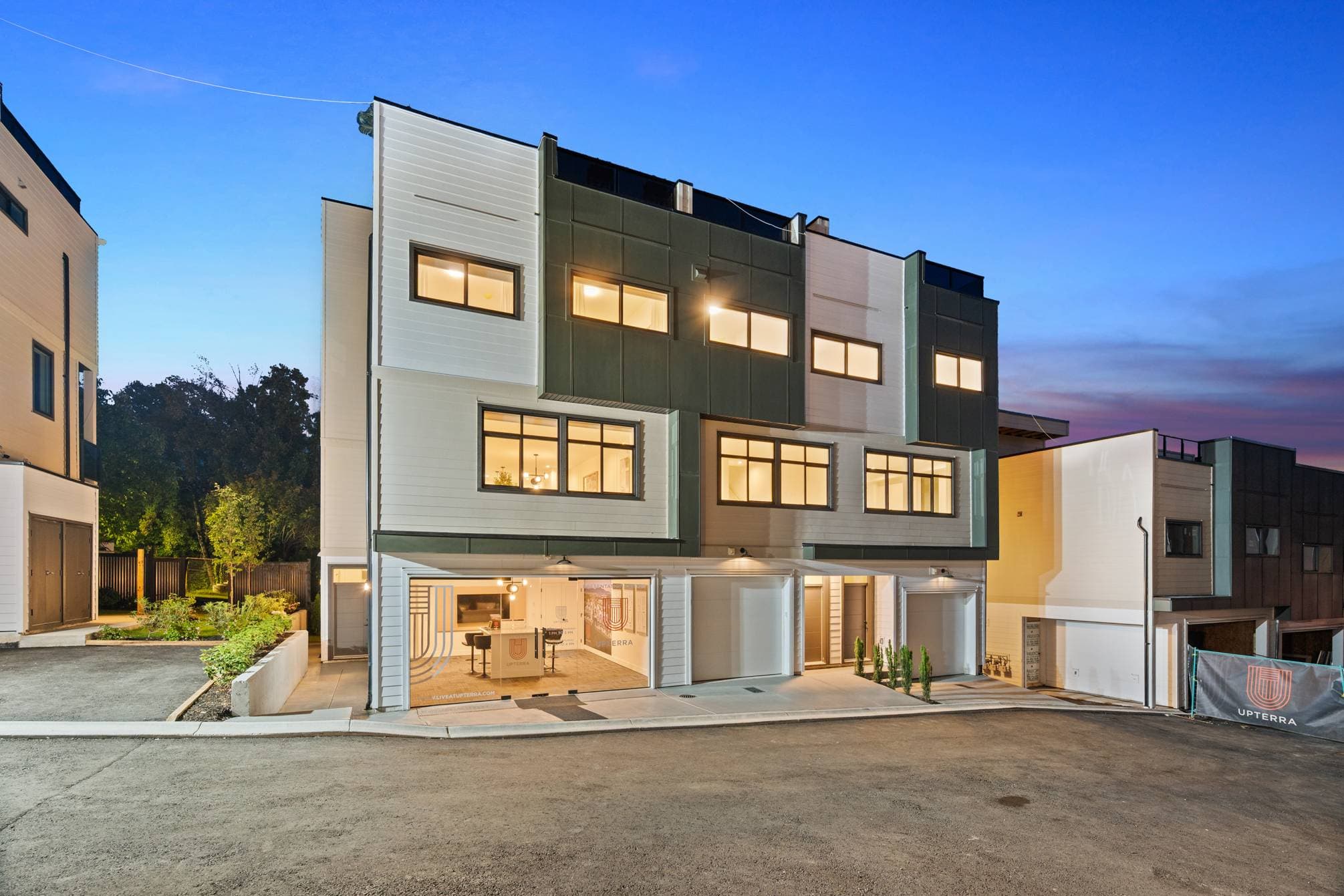 Upterra – A ground-level twilight view of a modern four-story townhouse complex featuring mixed light siding, dark green paneling, and glowing interior lights. One ground-floor garage bay is open and transformed into a sales center, facing a newly paved asphalt driveway under a clear dusk sky.