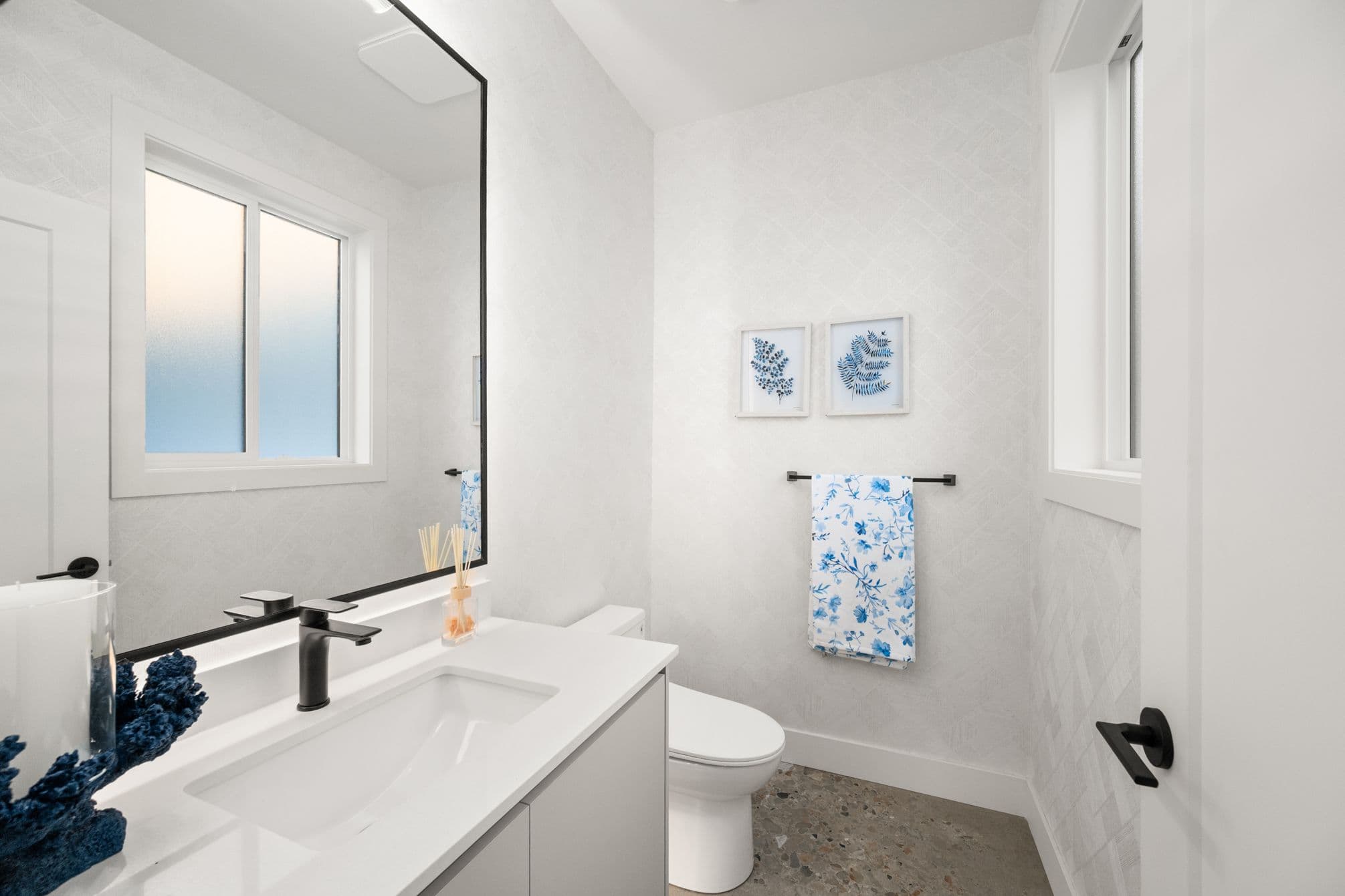 Upterra – A modern bathroom features a white vanity with a sleek black faucet, a large black-framed mirror, and a white toilet against a subtle textured wallpaper. Natural light enters through frosted windows, highlighting blue botanical artwork, a floral towel on a black rack, and polished terrazzo tile flooring.
