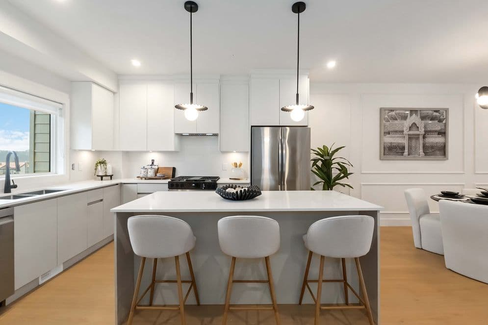 Upterra – A modern open-concept kitchen featuring a central island with a white quartz countertop, three white barstools with wooden legs, and two pendant lights hanging overhead. White flat-panel cabinetry, stainless steel appliances, light wood flooring, and an adjacent dining area complete the bright and clean space.