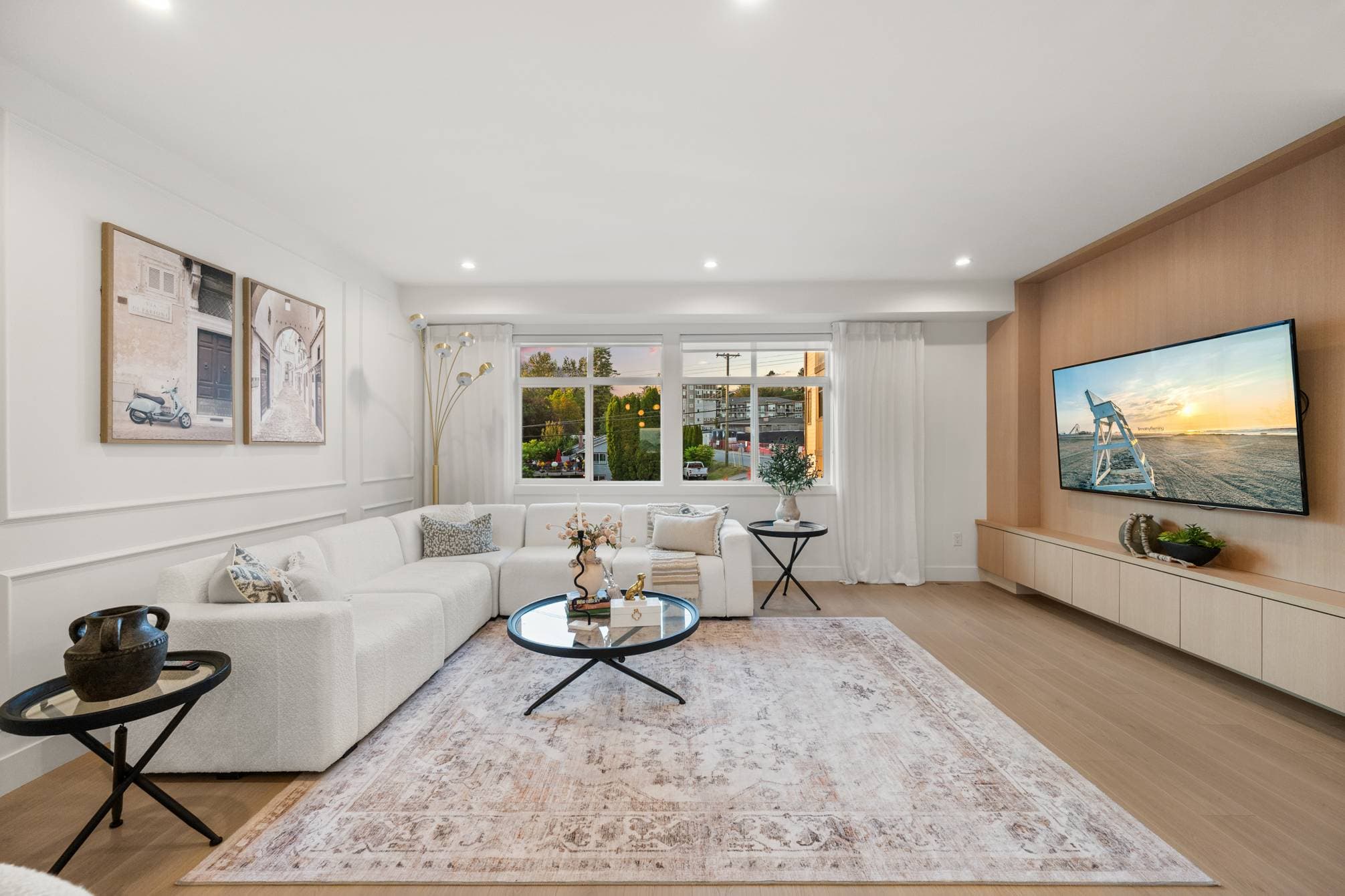 Upterra – A bright, modern living room features a large cream sectional sofa paired with a vintage-style pattern rug and a glass coffee table. A wall-mounted television is set into a custom wood accent wall with low cabinetry, complemented by light wood flooring and large windows letting in natural light.