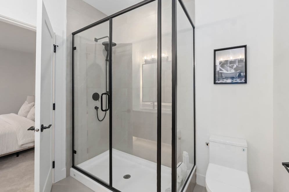 Upterra – A modern bathroom featuring a walk-in glass shower enclosure with bold black framing and hardware alongside a white toilet. Through an open white door to the left, a bright bedroom with a neatly made bed is visible.