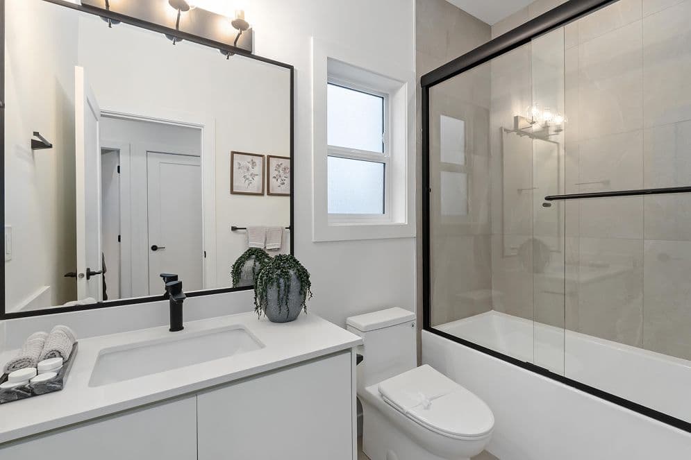 Upterra – A modern bathroom featuring a white vanity with matte black fixtures, a large black-framed mirror, and a toilet beside a glass-enclosed bathtub shower with neutral tiled walls. Bright daytime lighting fills the space from a frosted window, enhancing the clean, minimalist aesthetic.