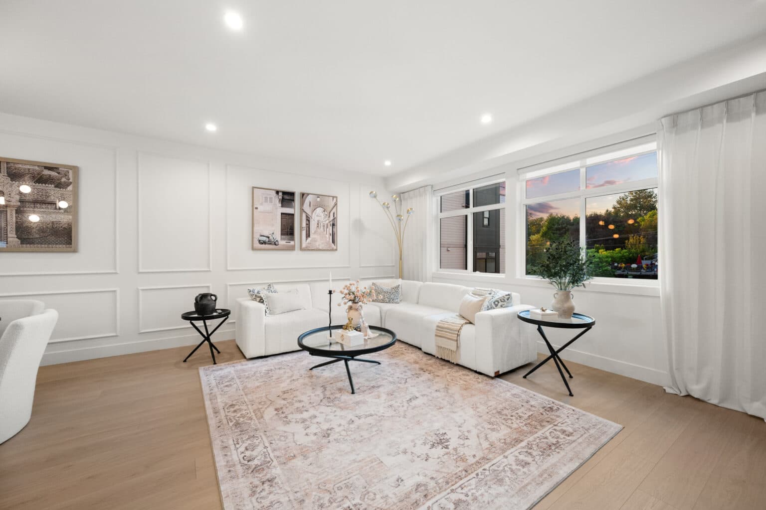 Upterra – A bright, modern living room featuring a white sectional sofa, light wood flooring, and detailed wainscoting paneling on the white walls. Large windows with sheer white drapes let in natural light, showcasing views of outdoor greenery and a soft sunset sky.