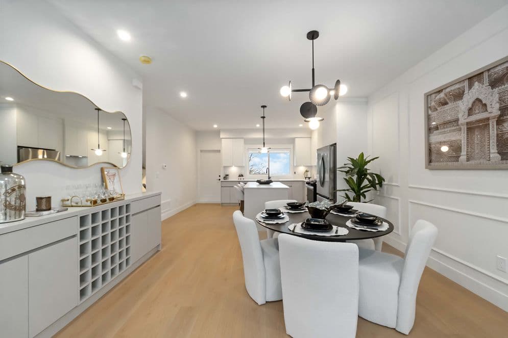 Upterra – An open-concept living area featuring a round dining table with dark place settings and white upholstered chairs, framed by a modern black sputnik chandelier. In the background, a bright kitchen with light grey cabinetry, stainless steel appliances, and a central island flows naturally into the brightly lit space with light hardwood floors.