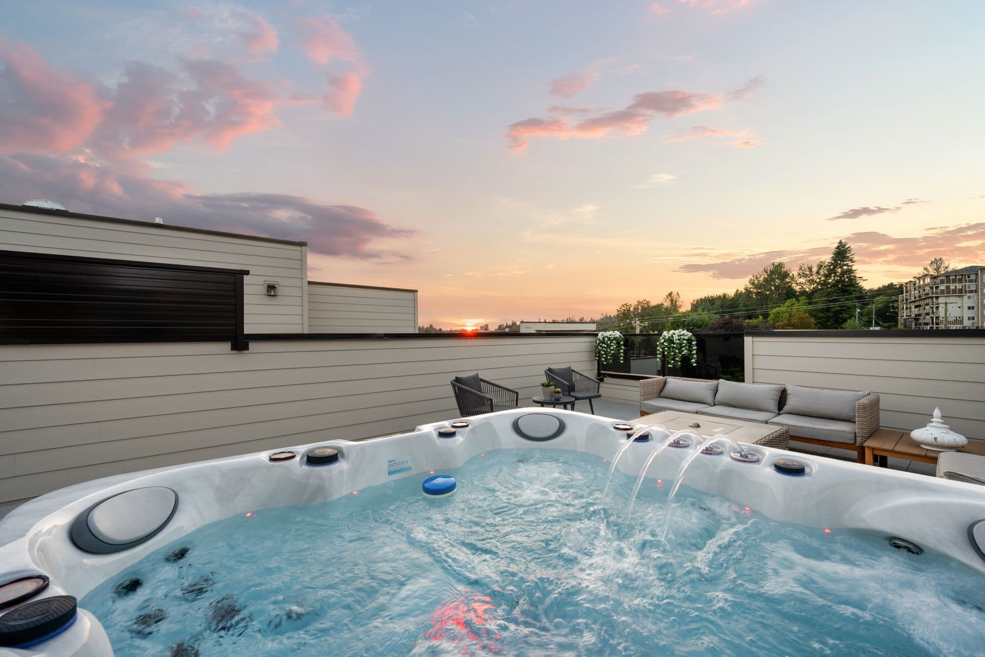 Upterra – A bubbling rooftop hot tub with water jets running takes center stage on a private terrace furnished with modern patio chairs and an outdoor sofa. The background showcases a vibrant sunset with pink and orange clouds stretching across the sky above distant treetops and neighboring residential buildings.