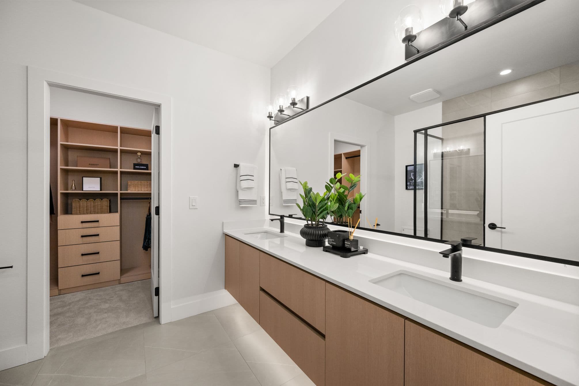 Upterra – A modern ensuite bathroom featuring a long floating light-wood double vanity with a white countertop, matte black hardware, and an expansive wall-to-wall mirror. An open doorway leads directly into a custom walk-in closet with built-in wood shelving and drawers.