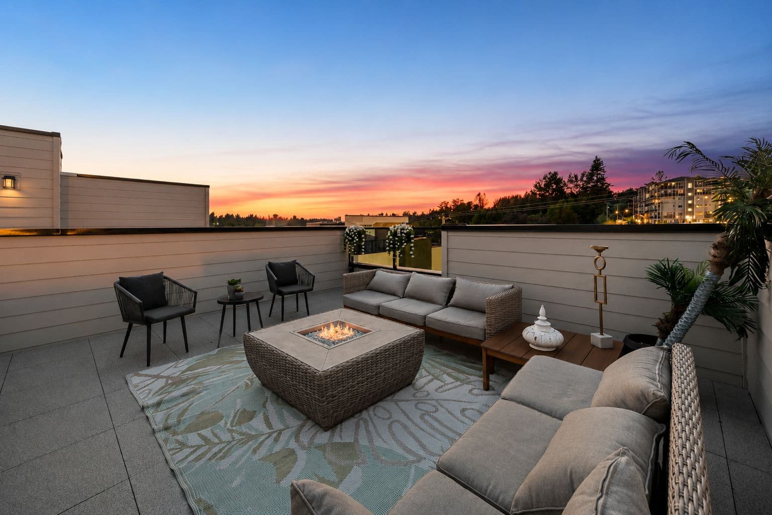 Upterra – A rooftop patio is furnished with woven outdoor sofas, black armchairs, an outdoor rug, and a central square fire pit with lit flames. The space overlooks distant trees and buildings during a vibrant dusk sky filled with pink, orange, and blue sunset colors.