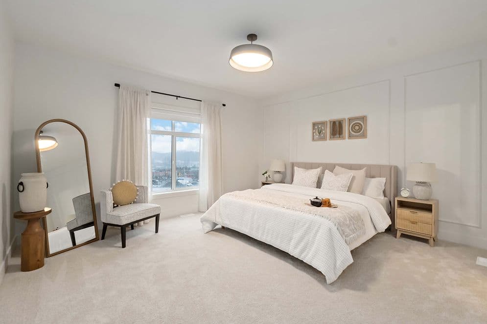 Upterra – A bright, neutral-toned bedroom features a king-sized bed dressed in white bedding, flanked by wooden nightstands and framed by wall molding. An arched full-length floor mirror and an accent chair sit beside a window that offers views of distant hills under a clear blue sky.
