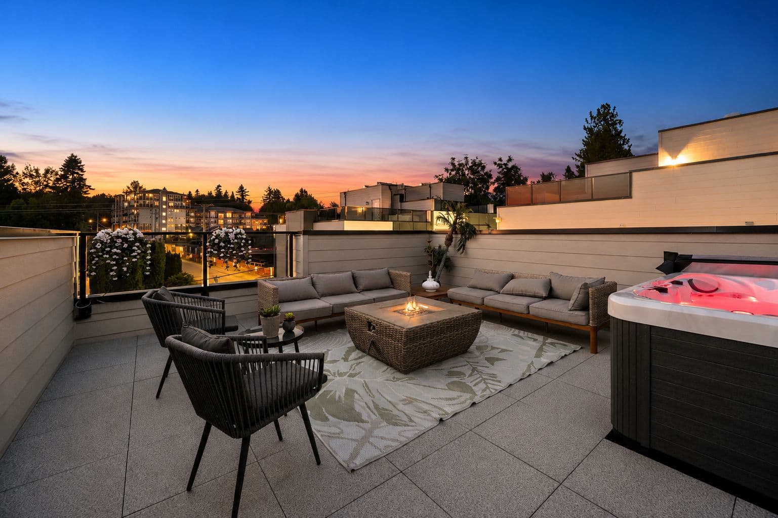 Upterra – A rooftop lounge terrace features cushioned patio seating arranged around a lit square fire table and an illuminated hot tub in the foreground. Glass railings offer open views of surrounding trees and distant buildings during a vibrant orange and blue dusk.