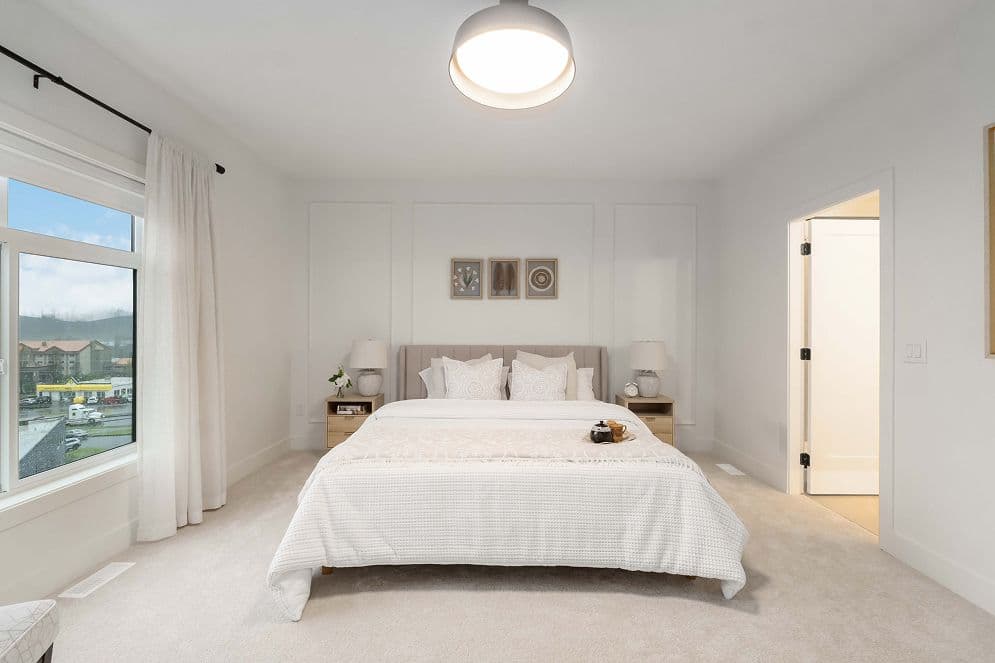 Upterra – A bright, modern bedroom featuring a neatly dressed king bed with a neutral, upholstered headboard and matching nightstands. Soft natural light streams in through a large window on the left, highlighting the clean white walls, wall molding accents, and plush beige carpeting.