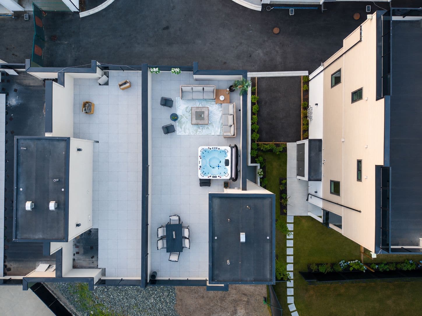 Upterra – A top-down aerial view showcases a modern residential rooftop deck outfitted with outdoor living furniture, including a hot tub, lounge sofas surrounding a coffee table, and an outdoor dining set. Adjacent paved pathways, dark rooftops, and light-colored building sidings frame the tiled patio space.