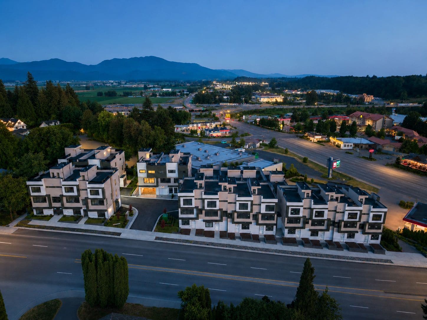 Upterra – An elevated twilight view of a modern multi-unit townhouse development situated along a multi-lane roadway. Brightly lit interiors glow from the contemporary geometric buildings, while sprawling farmland, rolling hills, and distant mountain silhouettes stretch under a deep blue sky.