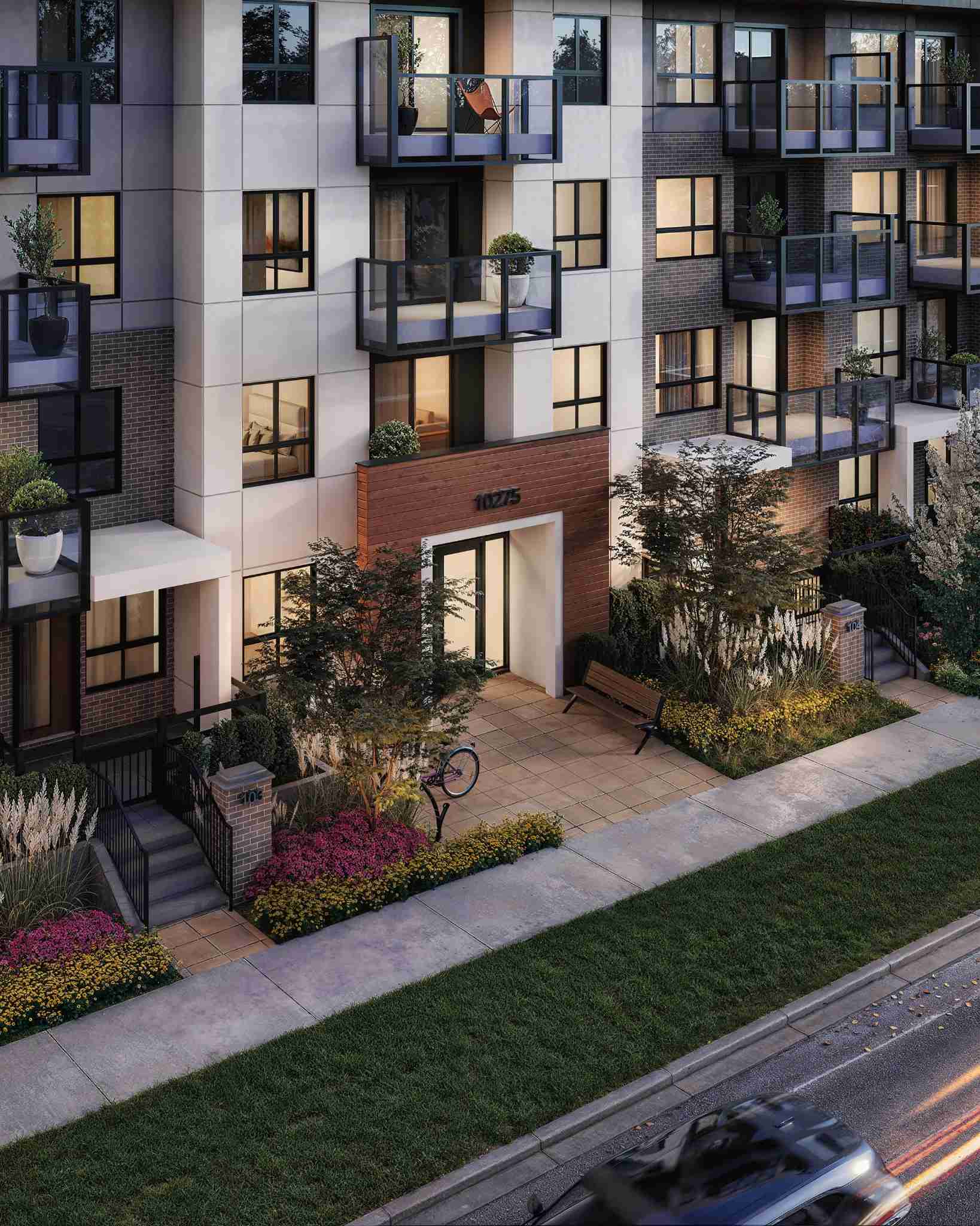 Muse – An elevated street-level view of a multi-story residential building facade clad in off-white panels and dark brick, featuring glass balconies and glowing interior windows. A wood-accented main entrance, numbered 10275, opens onto a paved forecourt with a bench, bicycle rack, and colorful landscaped flowerbeds bordering the sidewalk and street.