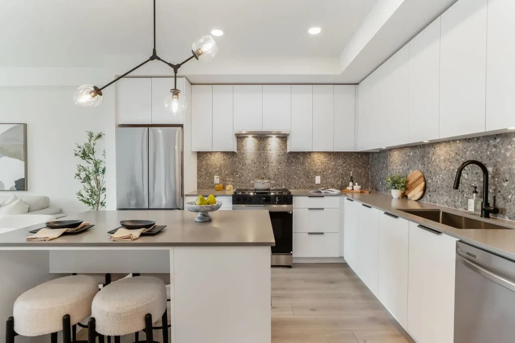 The Hive 2 – White kitchen with a stainless refrigerator, terrazzo-look backsplash, dark countertops, and an island with three cream stools.