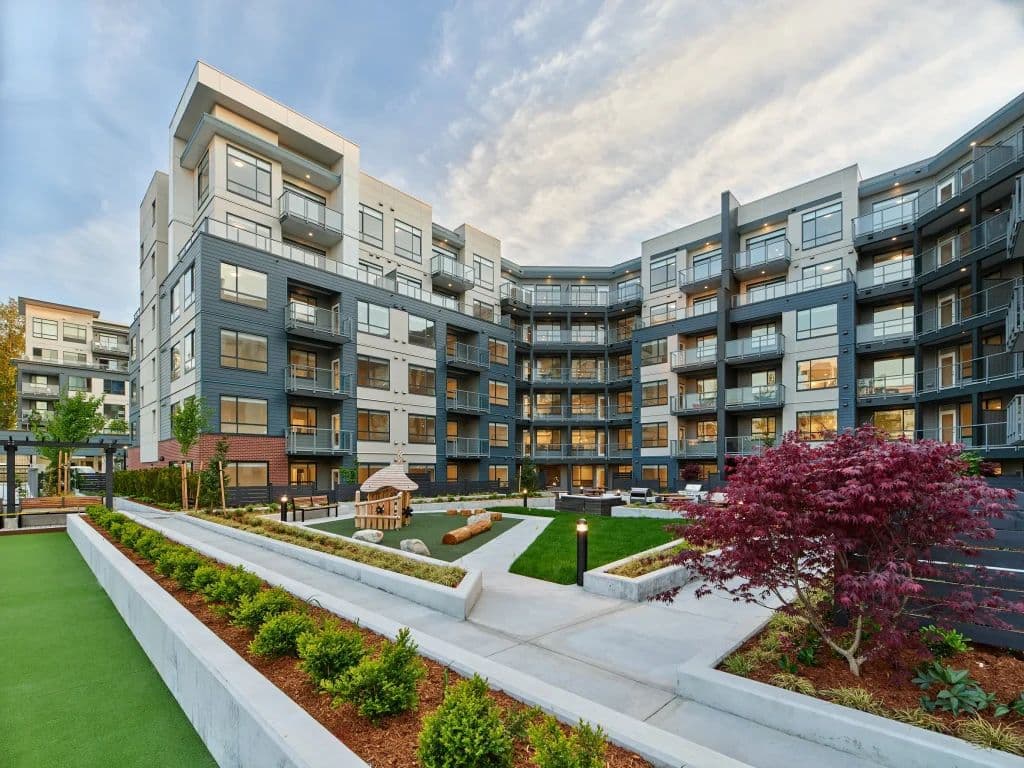The Hive 2 – Evening view of the building's interior courtyard with a children's play area, landscaped planters, and a maple tree, surrounded by lit balconies.