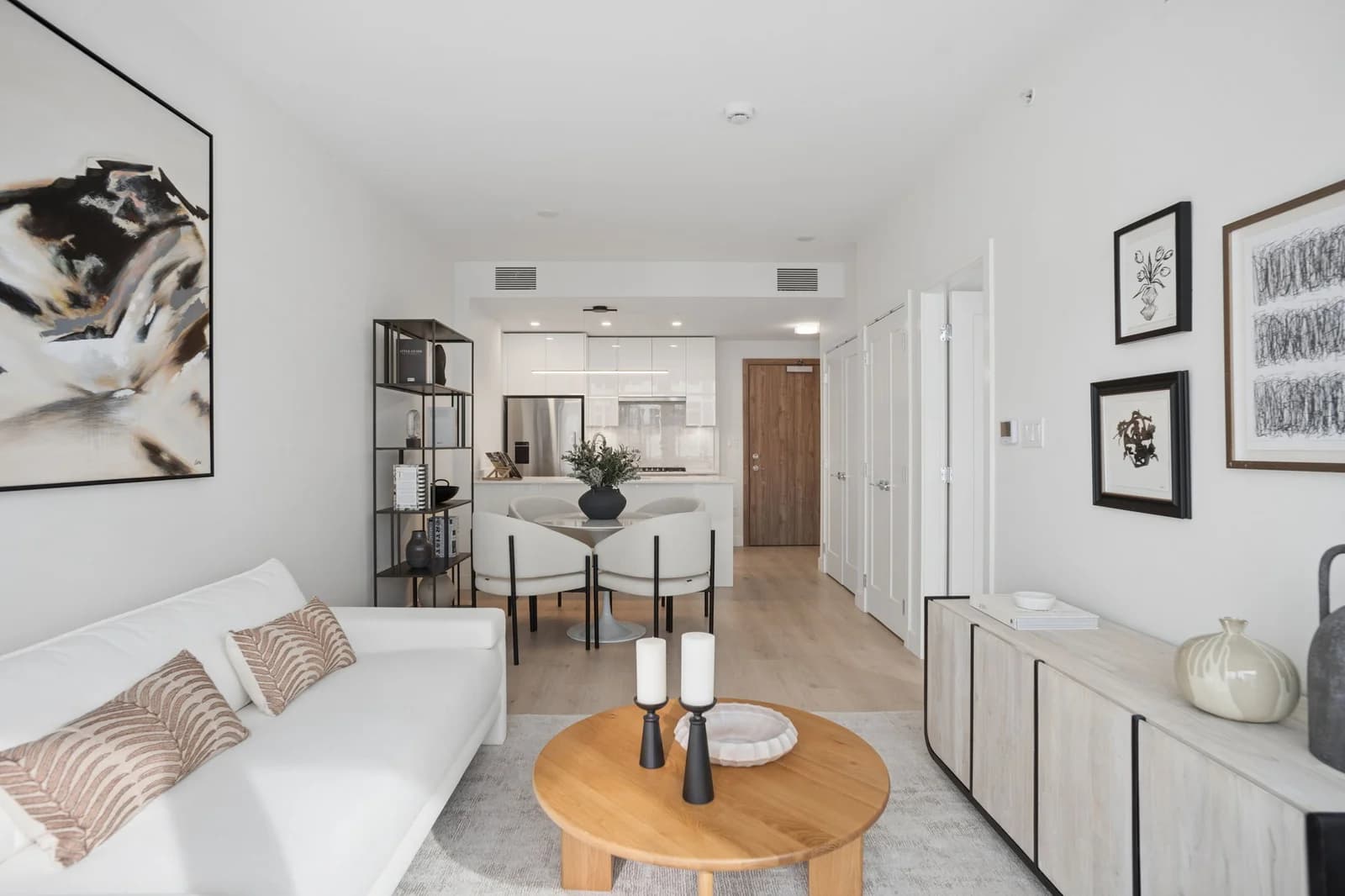 Milano at Capstan Village – A bright, open-concept living area features a white fabric sofa, a round wooden coffee table, and light wood flooring extending toward a small dining setup and a modern kitchen in the background. Clean white walls are accented with framed abstract artwork, minimalist black metal shelving, and a light-toned sideboard under soft indoor lighting.