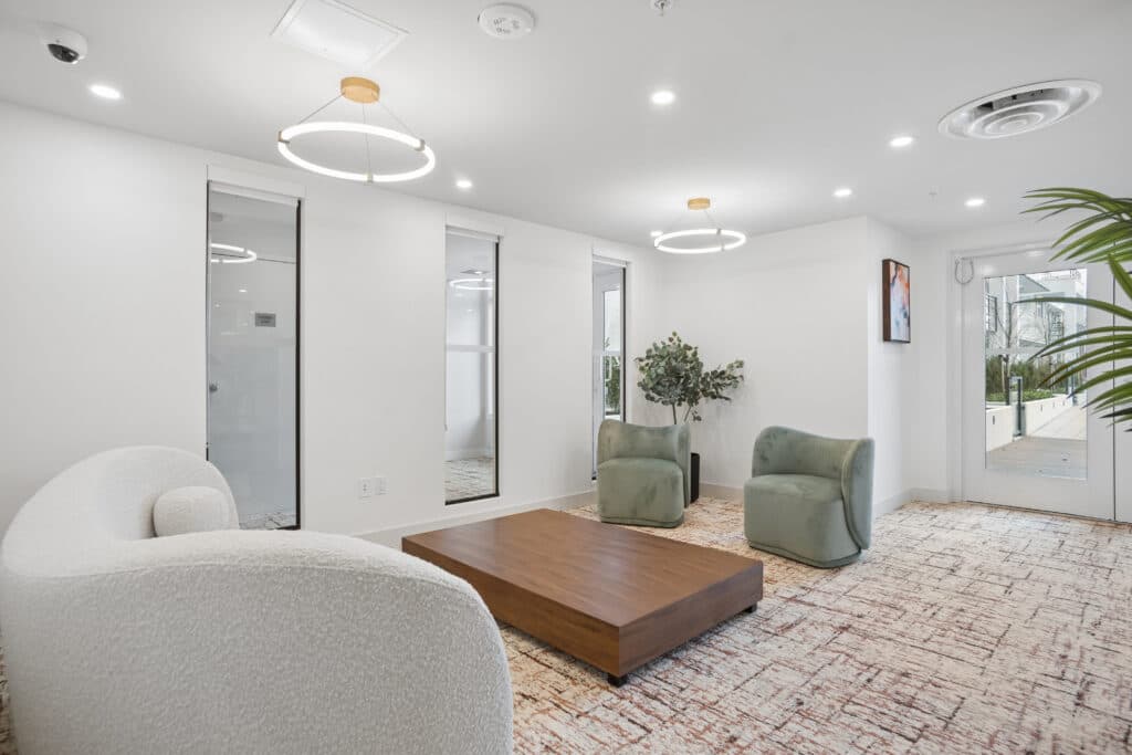 Milano at Capstan Village – A modern residential lounge feature a curved white bouclé sofa, two sage green armchairs, and a low wood coffee table set on a patterned area rug. White walls, recessed lighting, circular pendant light fixtures, and potted plants create a bright, contemporary atmosphere.