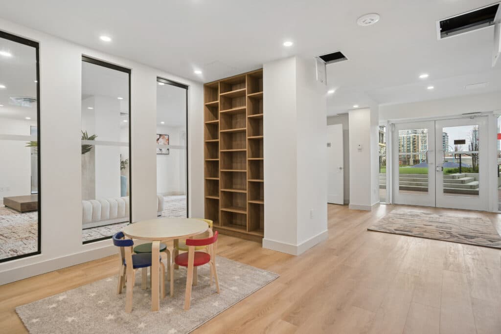 Milano at Capstan Village – A bright indoor play area features a small wooden children's table with colourful chairs set on a star-patterned rug over light-wood flooring. Tall floor-to-ceiling windows, built-in wooden shelving, and glass exit doors looking out to a courtyard create a clean, modern atmosphere.