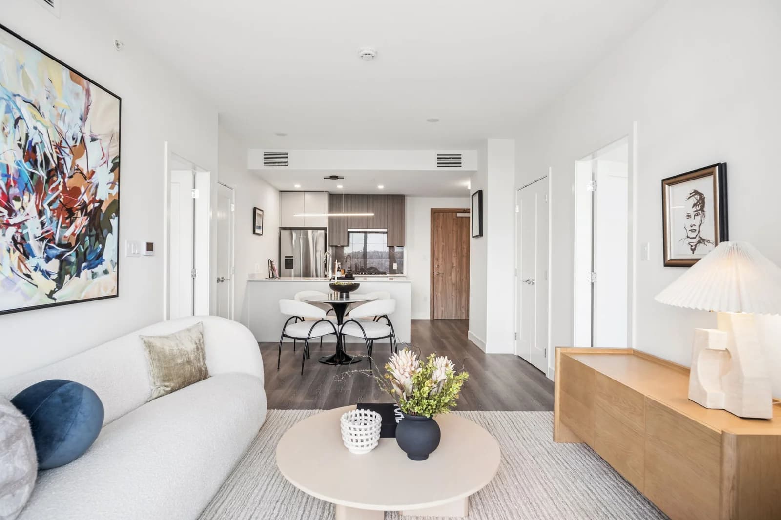 Milano at Capstan Village – A bright, open-concept living area features a white bouclé curved sofa, a round coffee table, and a wooden credenza under a sculpted lamp. Beyond the seating space sits a small round dining table leading into a contemporary kitchen with modern cabinetry and stainless steel appliances.