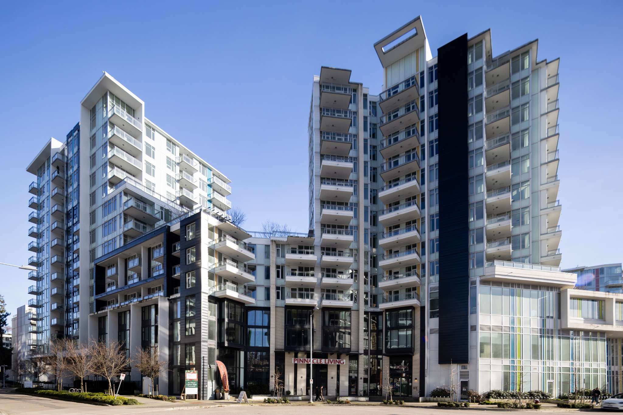 Milano at Capstan Village – A ground-level, street-side view shows a modern multi-story residential complex featuring glass balconies, white facades, and dark accent paneling under a clear blue sky. The building sits alongside a paved sidewalk with small street trees and a commercial entrance labeled 'Pinnacle Living' at the base.