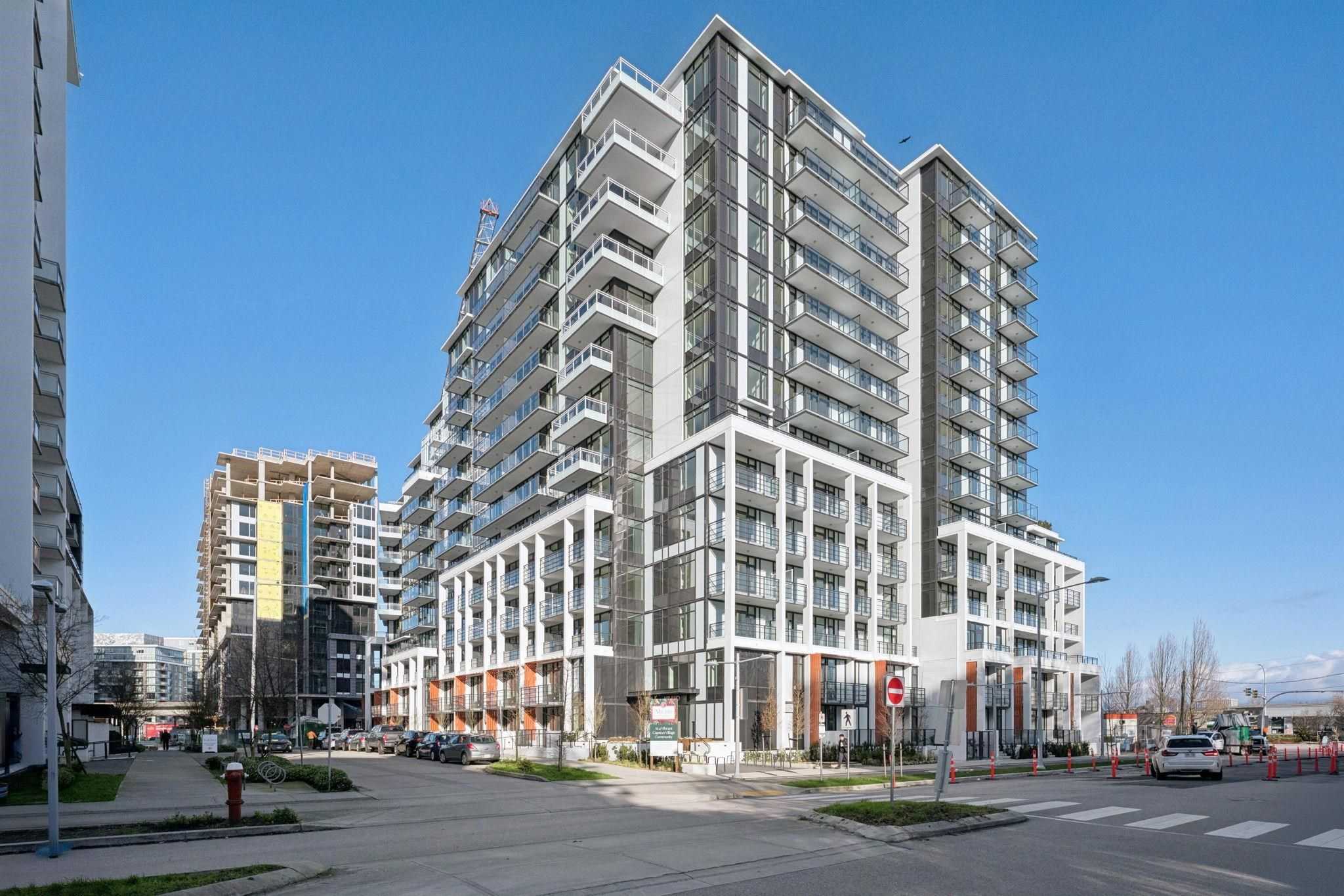 Milano at Capstan Village – A modern mid-rise residential tower with a white grid facade, glass balconies, and dark cladding stands on a sunny street corner under a clear blue sky. In the background, another mid-rise building is actively under construction alongside street-level sidewalks and parked cars.