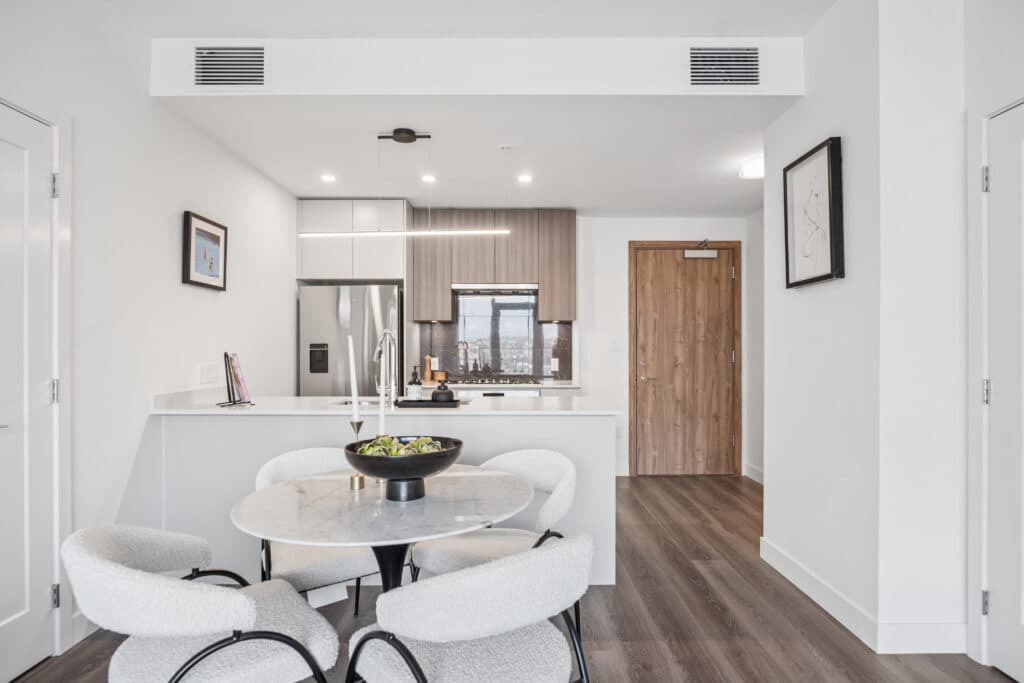Milano at Capstan Village – A modern open-concept dining and kitchen area features a round marble dining table with four textured white armchairs in the foreground. In the background, the kitchen offers flat-panel wood and white cabinetry, quartz countertops, stainless steel appliances, and dark wood-look laminate flooring under bright overhead lighting.
