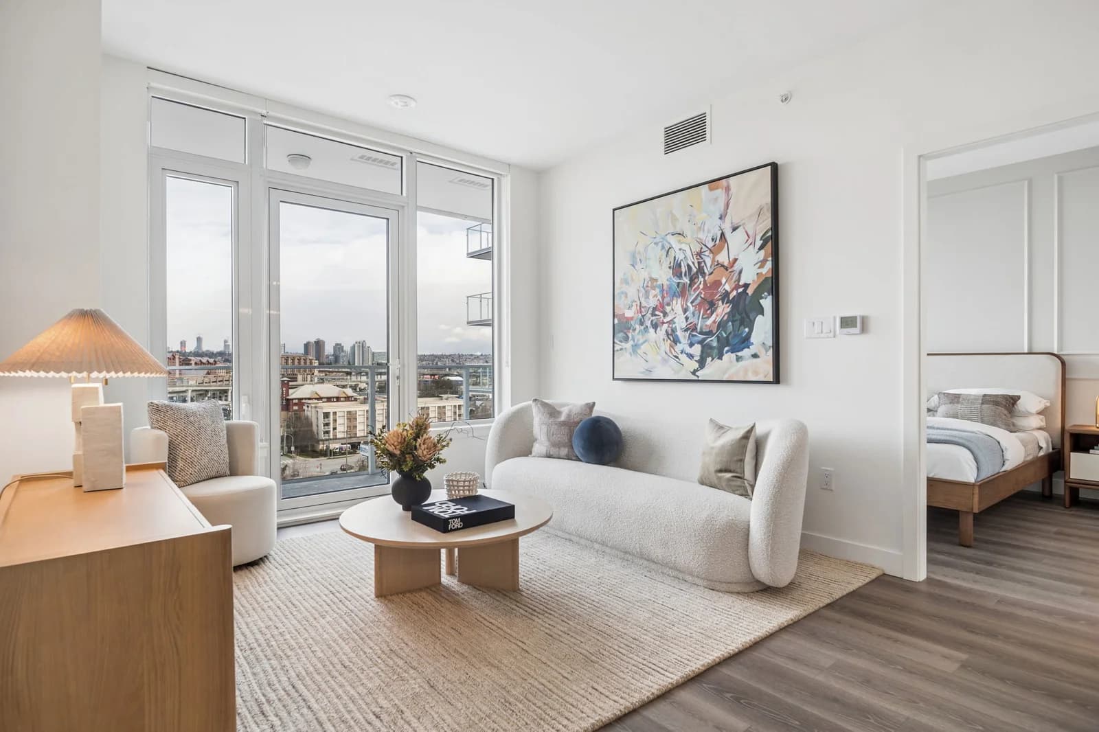 Milano at Capstan Village – A modern living room features a white bouclé curved sofa, a round wooden coffee table on a textured area rug, and floor-to-ceiling glass patio doors revealing a city view. To the side, an open doorway leads into a neatly styled bedroom with a wooden bed frame and neutral bedding.