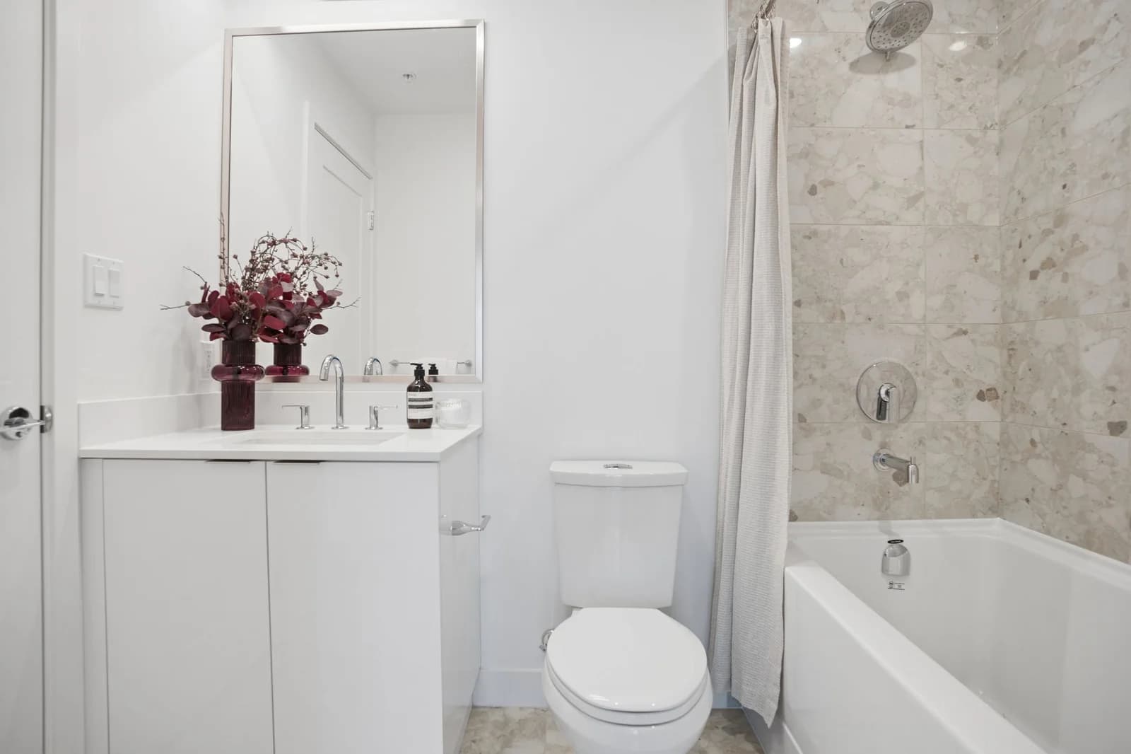 Milano at Capstan Village – A modern, bright bathroom featuring a white vanity with chrome hardware, a white toilet, and a bathtub surrounded by light beige terrazzo-style tiles. A deep red floral arrangement sits on the countertop next to a rectangular mirror, illuminated by clean bright indoor lighting.