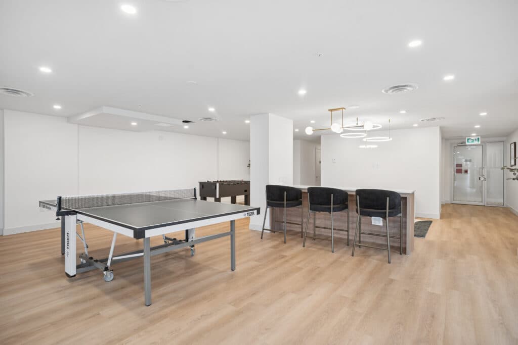 Milano at Capstan Village – A modern indoor game room and lounge featuring light wood-look flooring, crisp white walls, and bright recessed lighting paired with a contemporary ring chandelier. The space includes a ping pong table in the foreground, a foosball table in the background, and a counter with three dark upholstered bar stools.