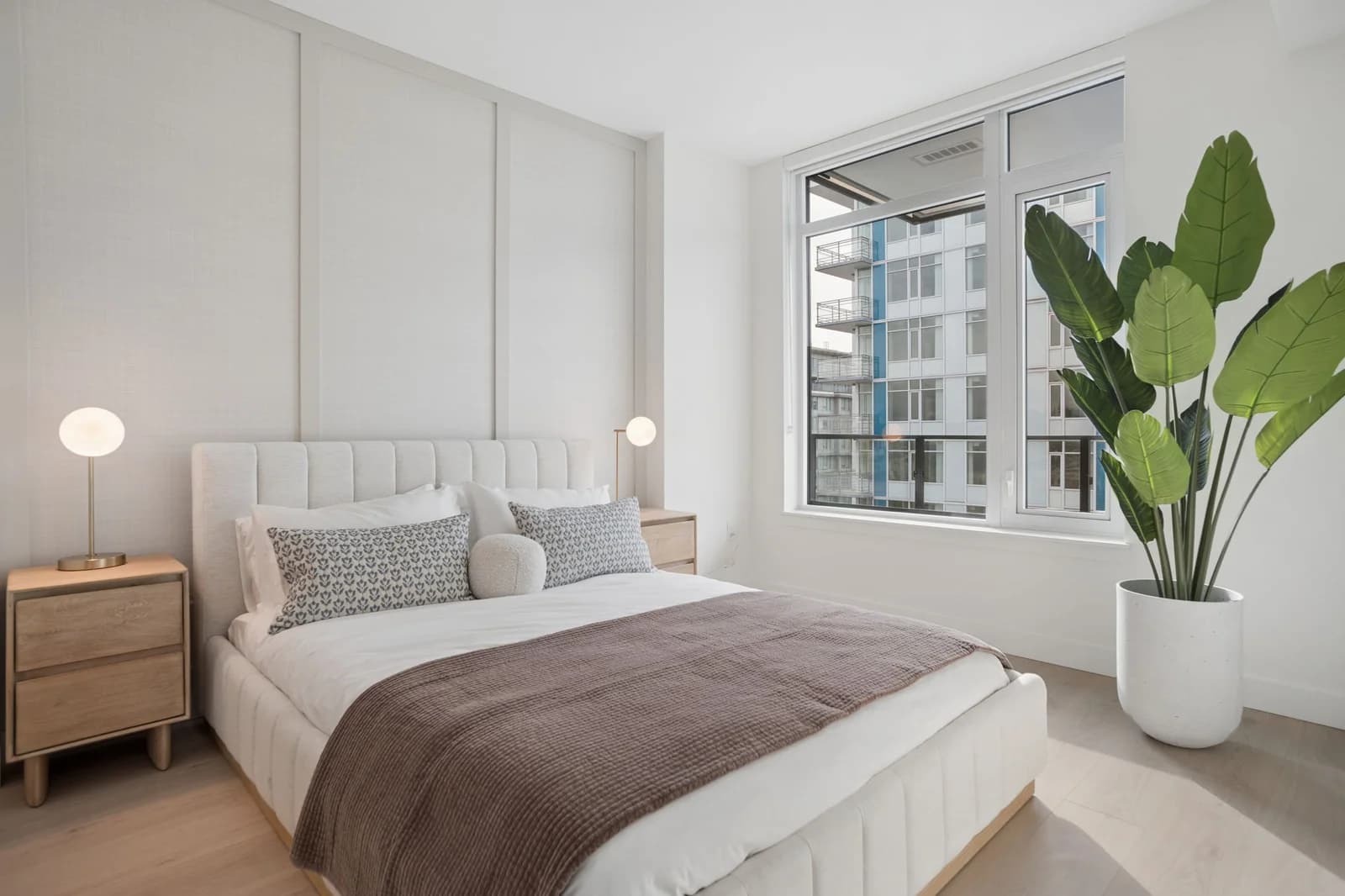 Milano at Capstan Village – A modern, bright bedroom features a tufted beige bed frame, white linens with a brown throw blanket, and matching light wood nightstands topped with spherical globe lamps. A large potted bird-of-paradise plant stands in the corner beside a large window that offers natural light and views of neighboring residential buildings.